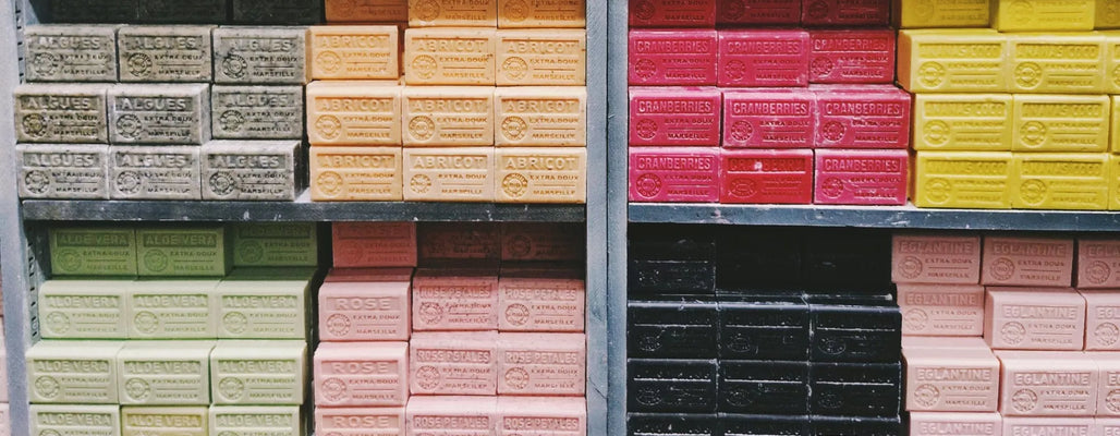Colorful artisan cold process soap bars stacked on retail shelves, organized by scent varieties including apricot, aloe vera, rose, cranberry, and coconut.