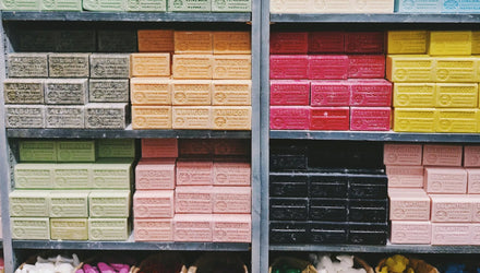 Colorful artisan cold process soap bars stacked on retail shelves, organized by scent varieties including apricot, aloe vera, rose, cranberry, and coconut.
